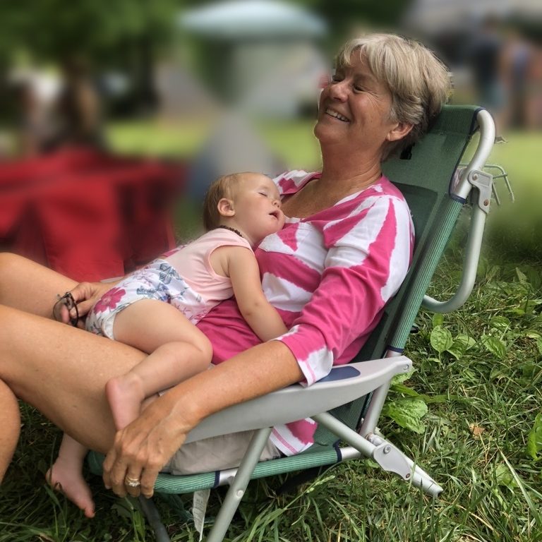 A smiling grandmother relaxing in a lawn chair at an outdoor market with her grandbaby asleep on her chest